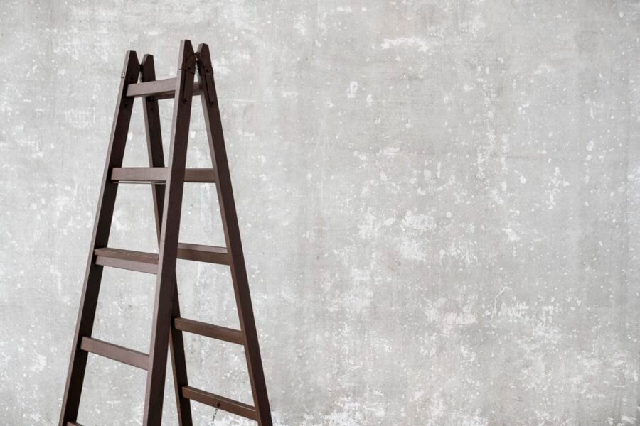 Scala a doppia salita in legno scuro posizionata contro una parete grigia con texture ruvida, ideale per lavori domestici o professionali.