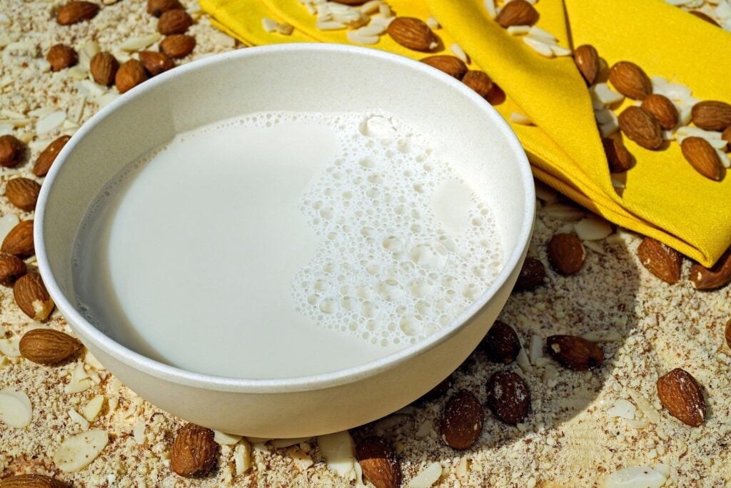 Ciotola di latte vegetale circondata da mandorle, con un tovagliolo giallo sullo sfondo.