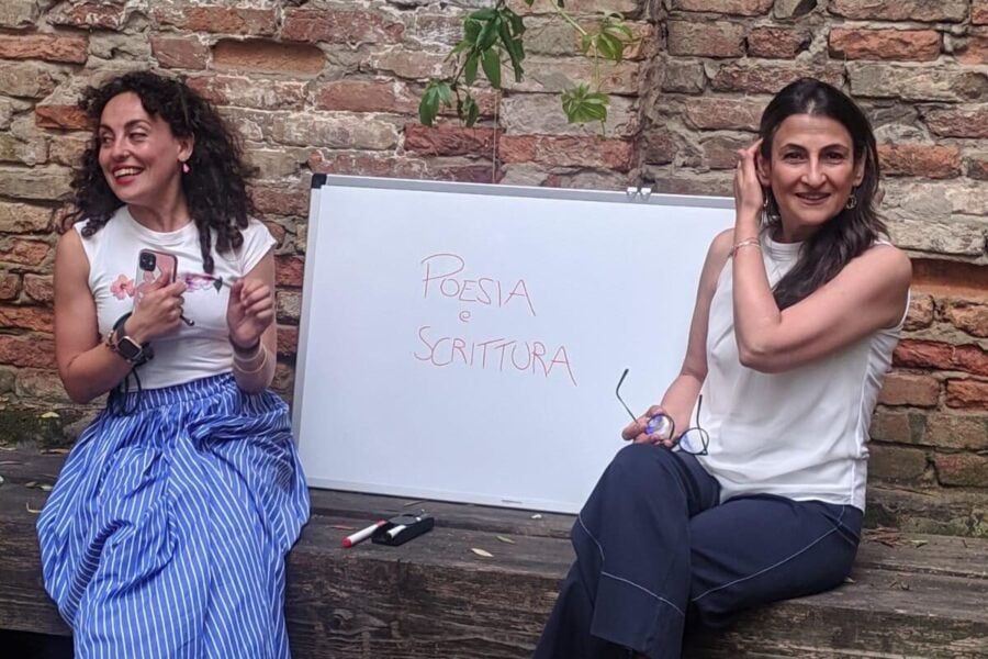 Salone Libro Torino 2025: in primo piano due ragazze sedute su una panchina, vicino a una lavagna con su scritto:" POESIA e SCRITTURA".